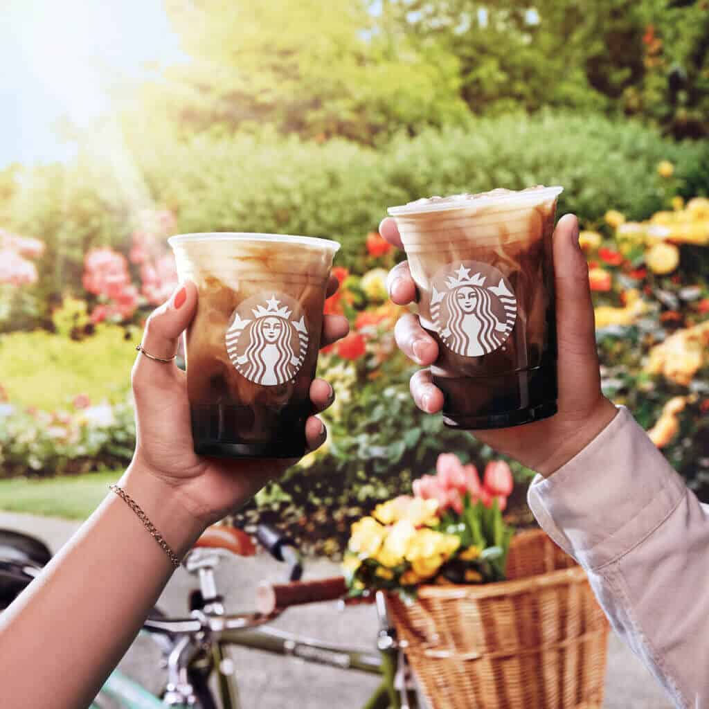 Two Iced Toasted Vanilla Oatmilk Shaken Espresso drinks held by hands in front of flower bushes.