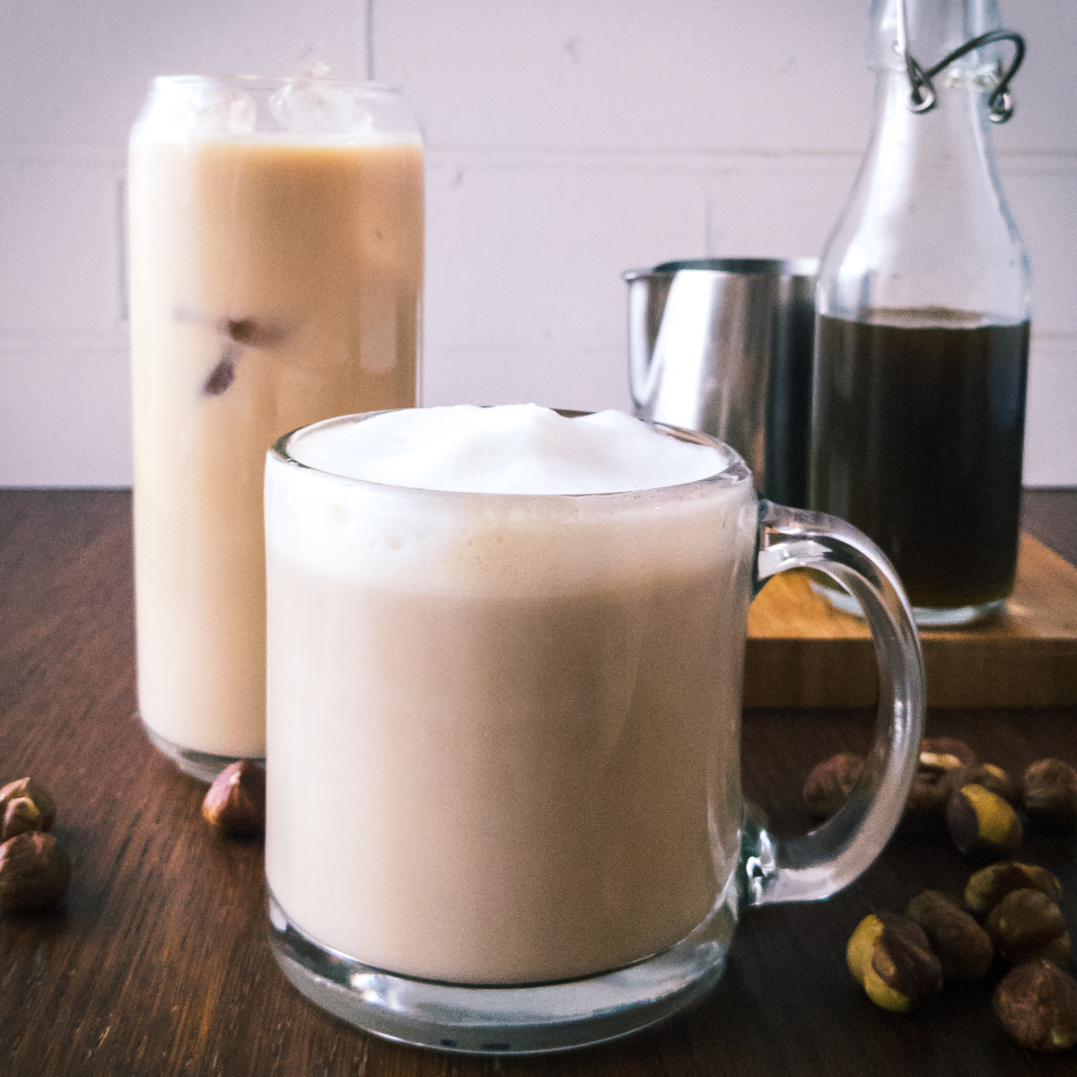 A hot and iced hazelnut latte sitting on a table with hazelnut syrup in the background.