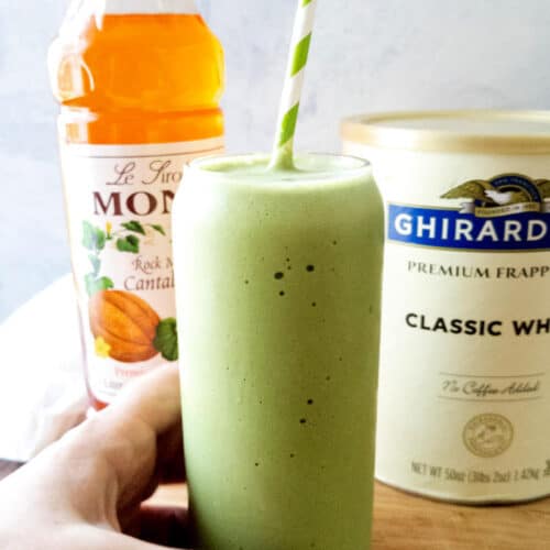 A matcha frappe in a clear glass with a hand holding it in the foreground with cantaloupe syrup and frappé mix in the background.